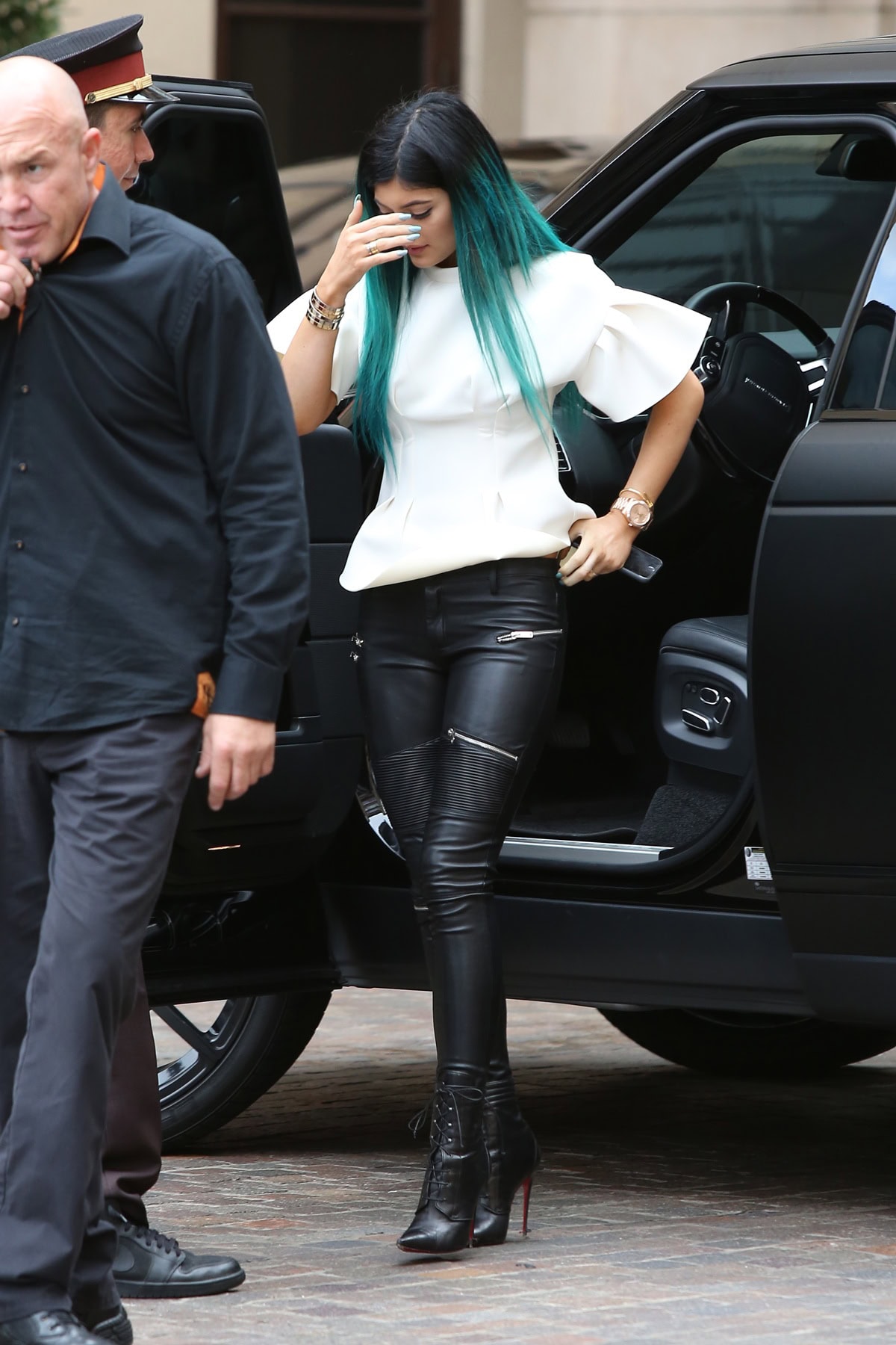 Kylie Jenner steps out of her black SUV, shielding her face from photographers as she arrives at Kourtney Kardashian’s baby shower on November 12, 2014, dressed in a white neoprene top, leather biker pants, and lace-up Christian Louboutin boots