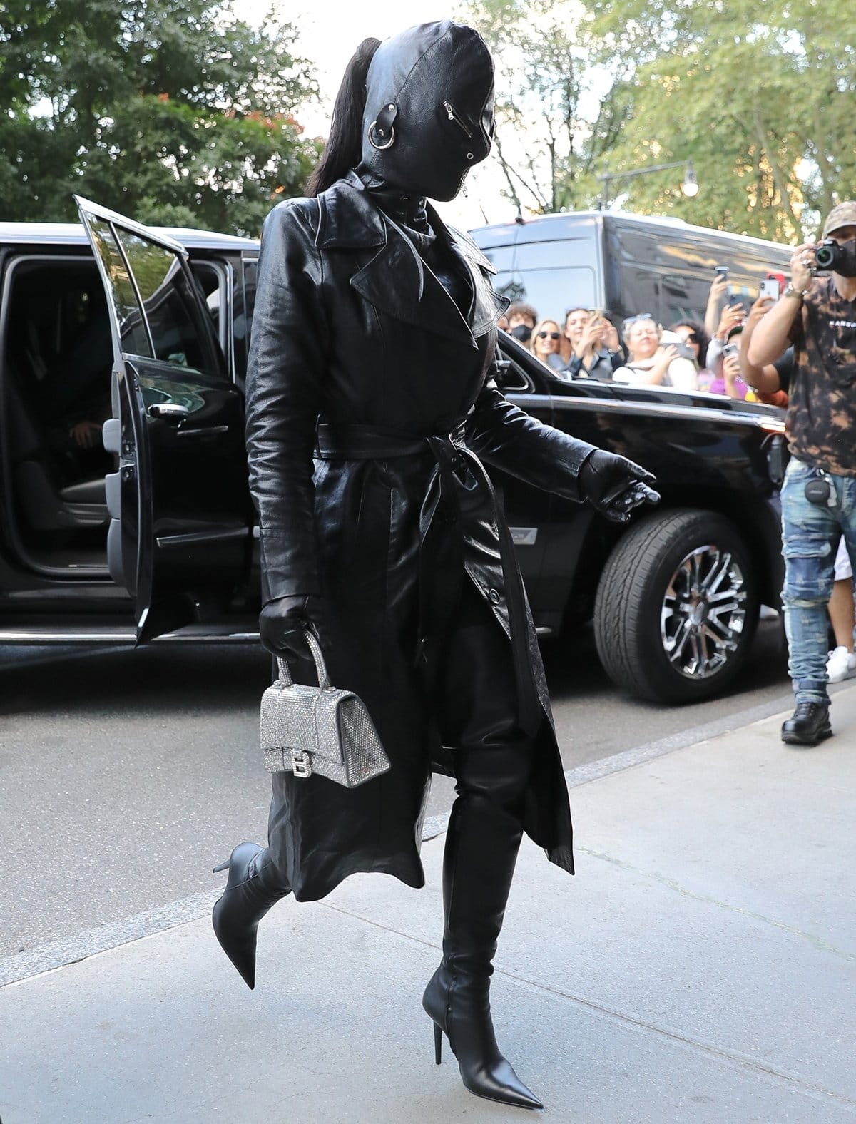 Kim Kardashian wears a black fetish mask with a matching leather trench coat, pants, and stiletto heel boots