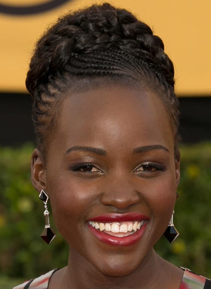 Lupita Nyong’o radiates with a braided updo, glowing skin, and vibrant red lipstick, complemented by her Art-Deco-inspired earrings at the 2015 SAG Awards