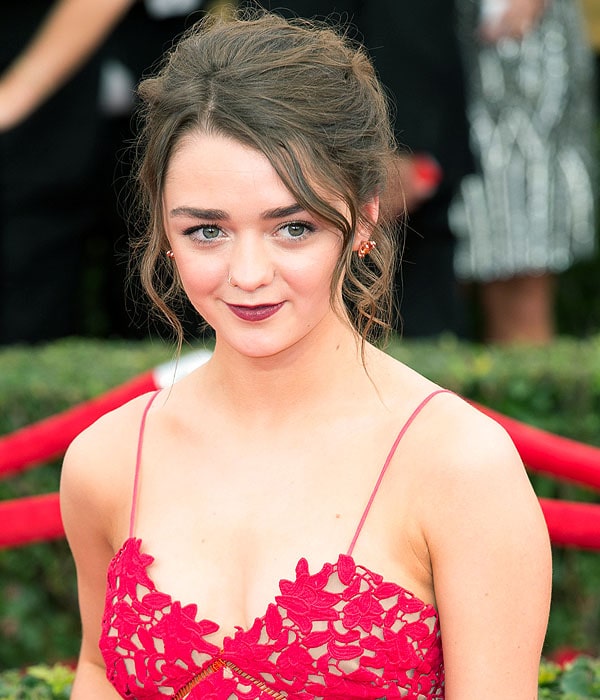 Maisie Williams wearing burgundy lips, red-jewel gold stud earrings, and a small hoop nose ring
