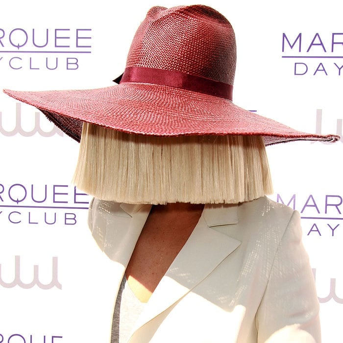 Sia hiding her face behind a bluntly cut blonde wig and a wide-brimmed maroon hat
