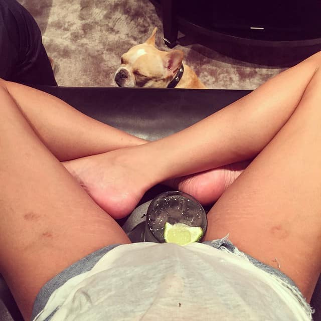 Chrissy Teigen relaxes with a drink on her lap, proudly displaying her stretch marks and legs in denim shorts, while her adorable dog sits contentedly in the background