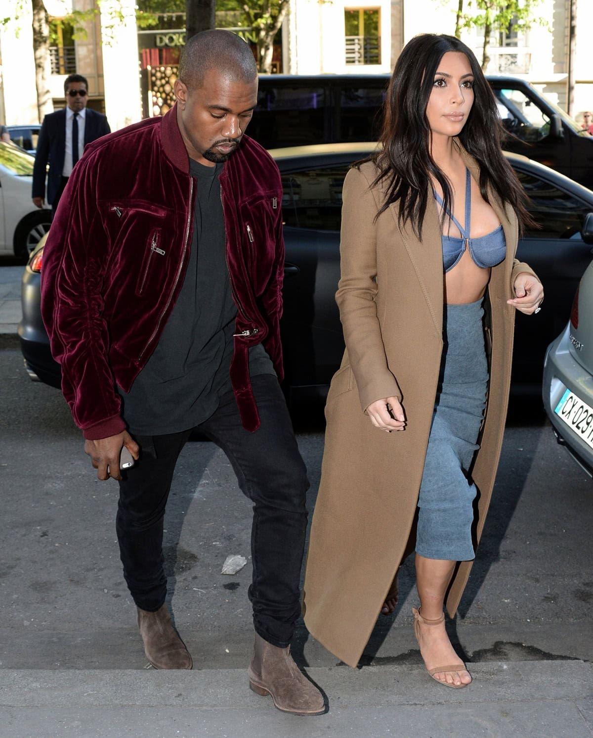 Kim Kardashian stuns in a denim bralette and suede pencil skirt paired with a camel coat and strappy heels, while Kanye West looks stylish in a burgundy velvet bomber jacket, black jeans, and suede Chelsea boots