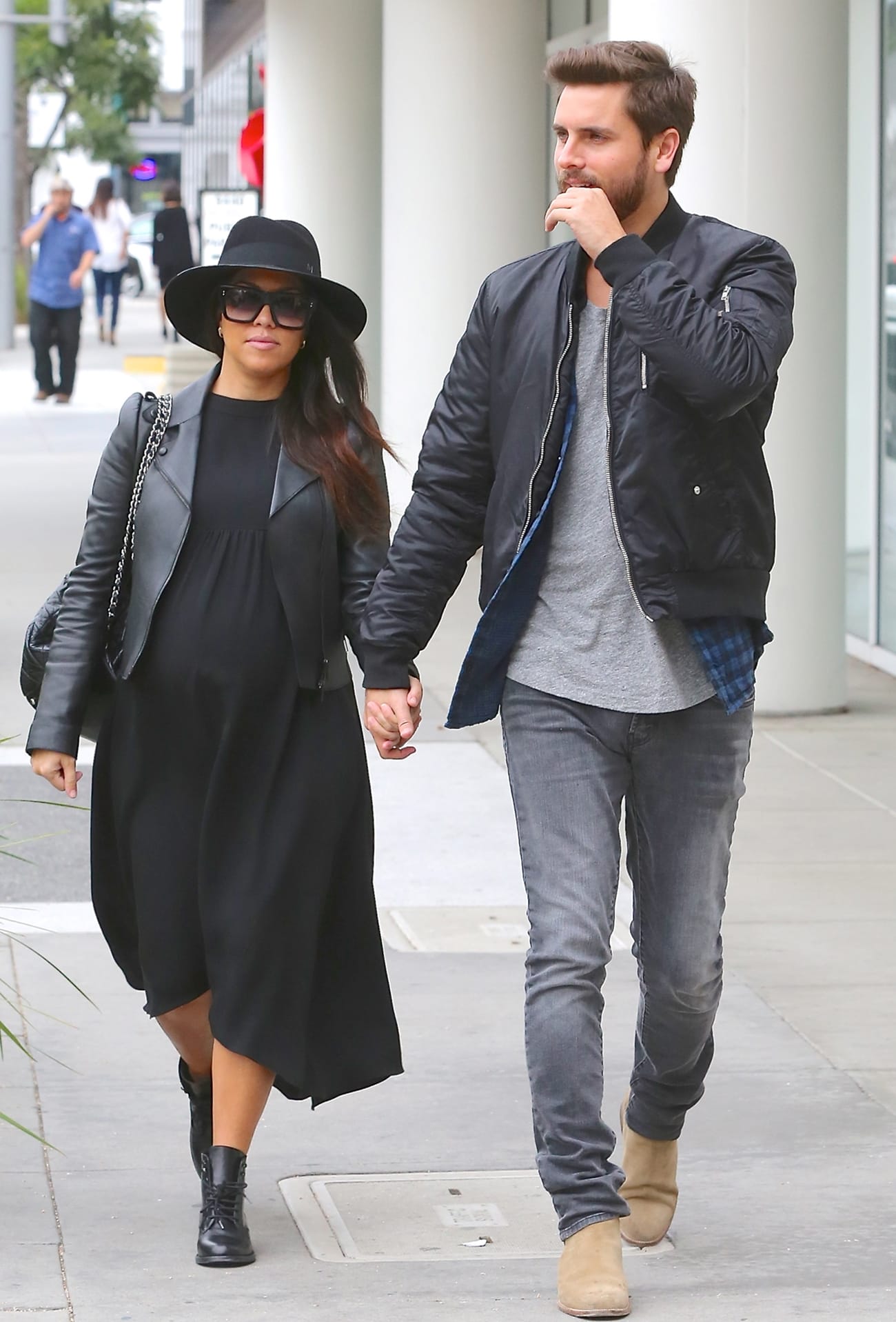 Kourtney Kardashian and Scott Disick step out in coordinated monochrome outfits, with Kourtney looking chic in a flowing black dress, leather jacket, wide-brim hat, and lace-up boots, while Scott keeps it casual in a bomber jacket, gray tee, skinny jeans, and suede Chelsea boots