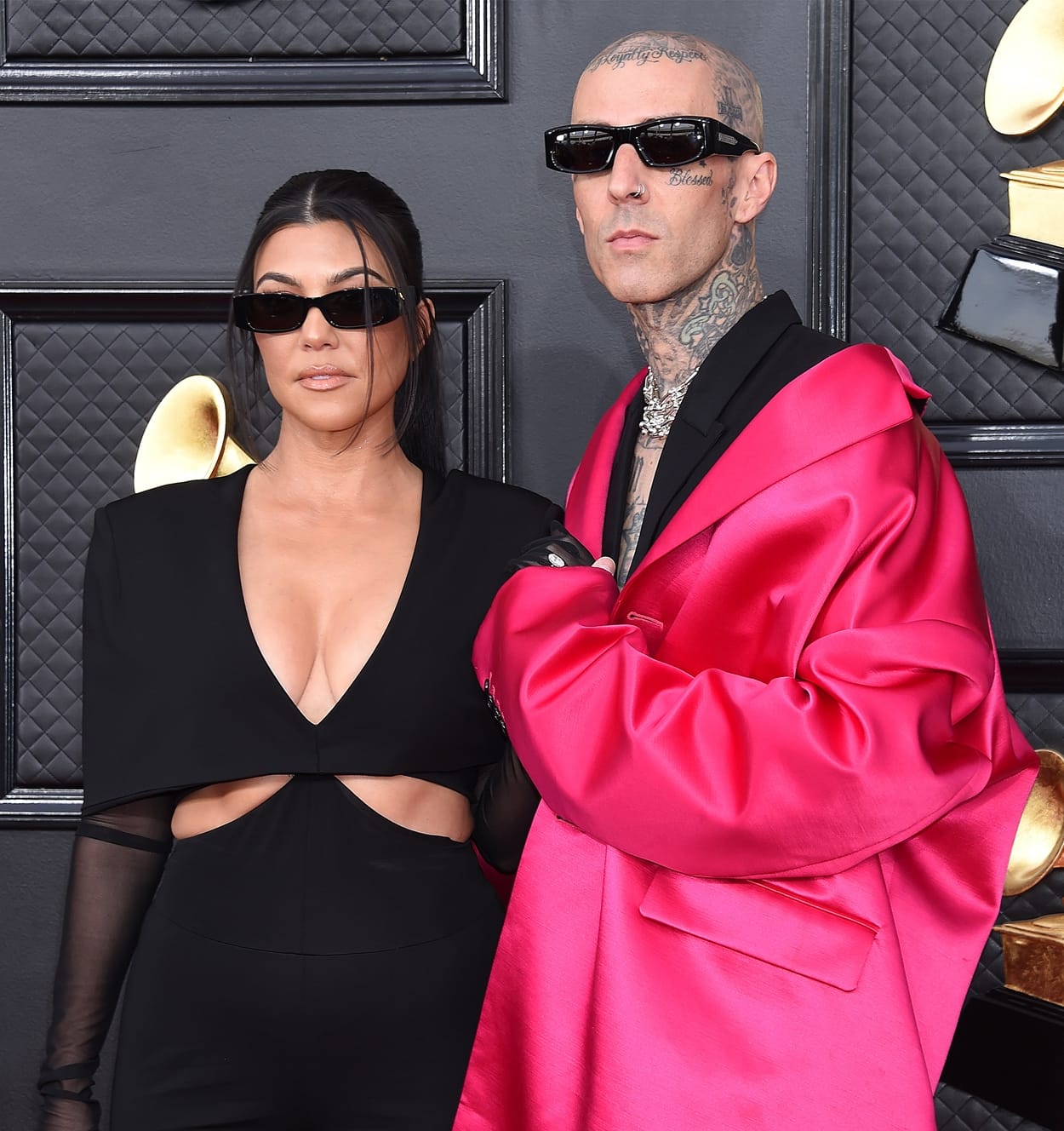 Kourtney Kardashian and Travis Barker make a bold fashion statement on the red carpet, with Kourtney in a sleek black cutout jumpsuit featuring sheer gloves and sharp sunglasses, while Travis contrasts in a striking oversized hot pink satin blazer, black outfit, and edgy accessories