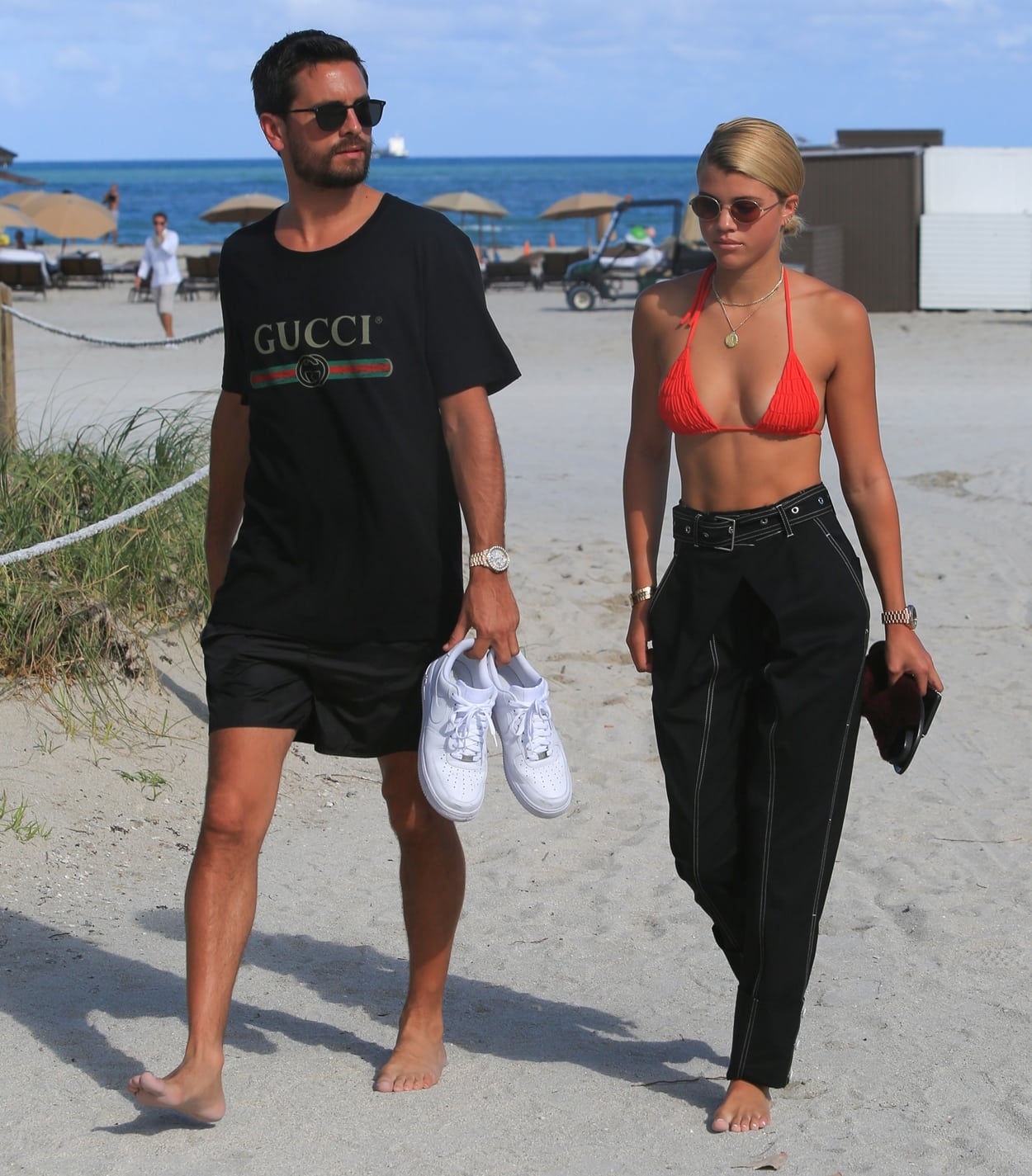 Scott Disick and Sofia Richie stroll along the beach in stylish yet contrasting outfits, with Scott in a relaxed black Gucci T-shirt, black shorts, and sunglasses while carrying his white sneakers, while Sofia stuns in a bold red bikini top paired with high-waisted black trousers, accessorized with gold jewelry and round sunglasses
