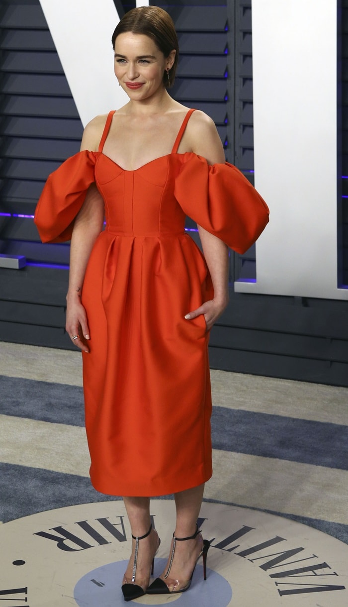 Emilia Clarke in a red Rosie Assoulin Fall 2019 dress with Christian Louboutin shoes, a Tyler Ellis clutch, Nak Armstrong earrings, and an Eriness Jewelry ring