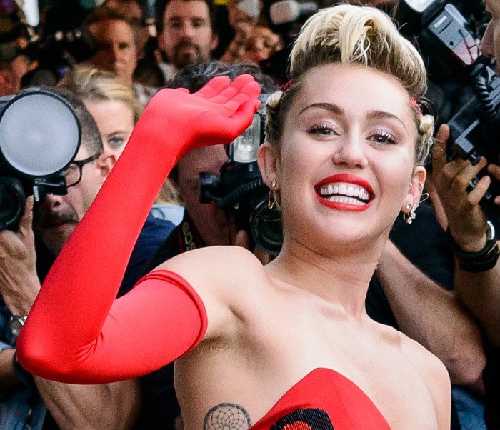 Miley Cyrus smiles in red, showing off her underarm hair and ribcage tattoo