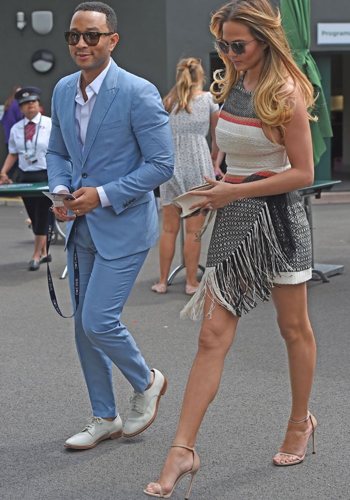 Chrissy Teigen adds a touch of elegance to her Wimbledon look, carrying a chic cream handbag with a gold chain strap that perfectly complements her fringed mini dress