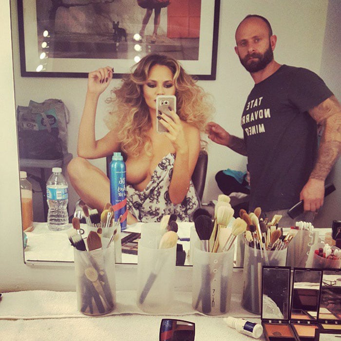 Chrissy Teigen taking a photo of her getting glammed up, with a bottle of hairspray covering her nipple