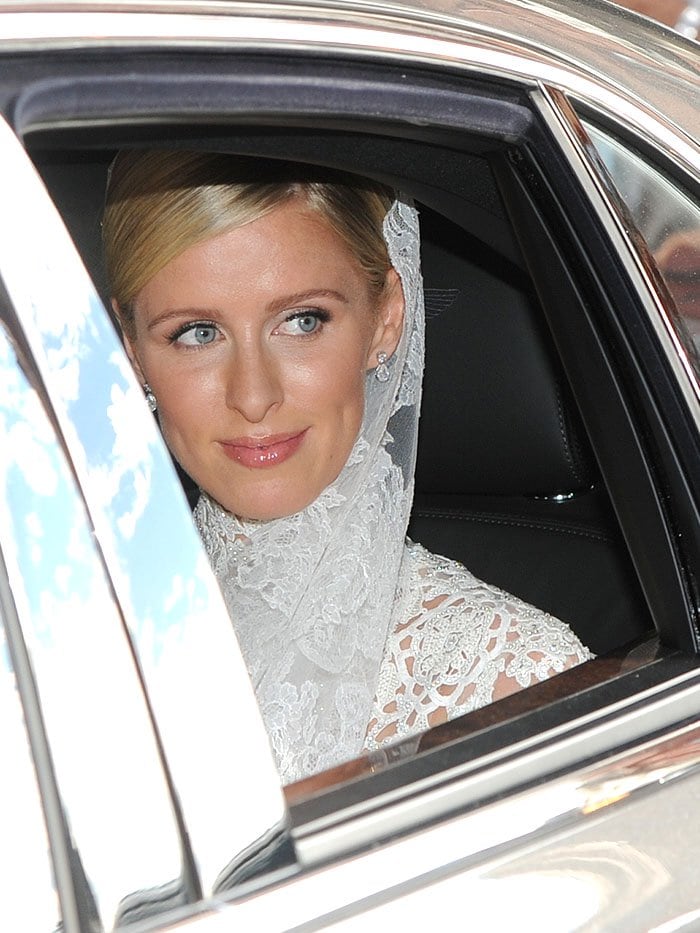 Nicky Hilton is seen sitting inside a luxury car, wearing a delicate lace veil that frames her face, showcasing her soft bridal makeup and elegant updo