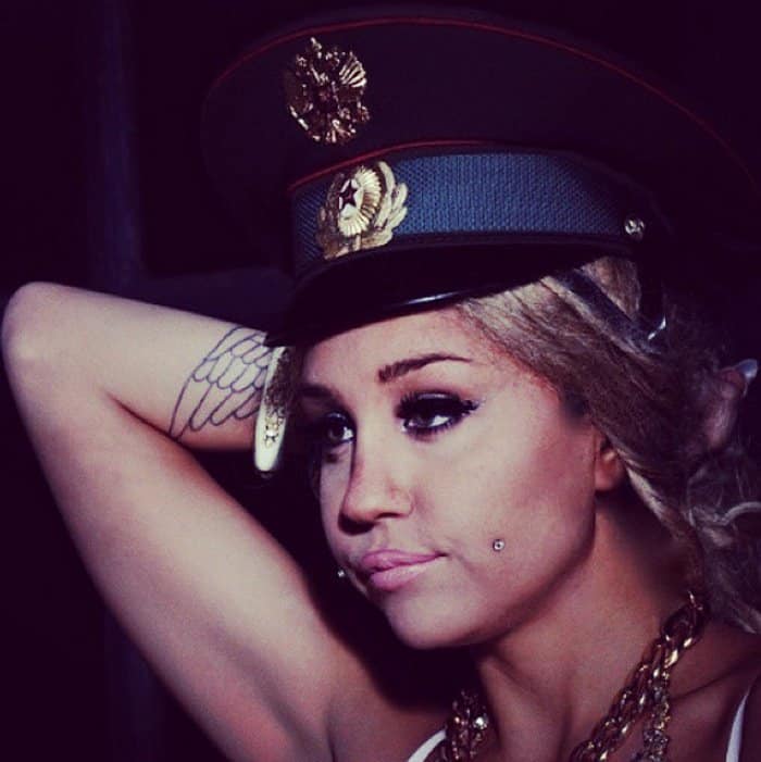 Amanda Bynes wearing a military-style hat, showcasing a cheek piercing and wing tattoo on her arm