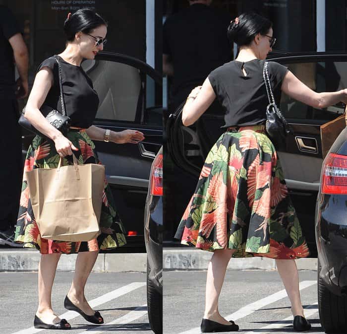 Dita Von Teese wears a vibrant, pleated midi skirt featuring a striking leaf and bird print