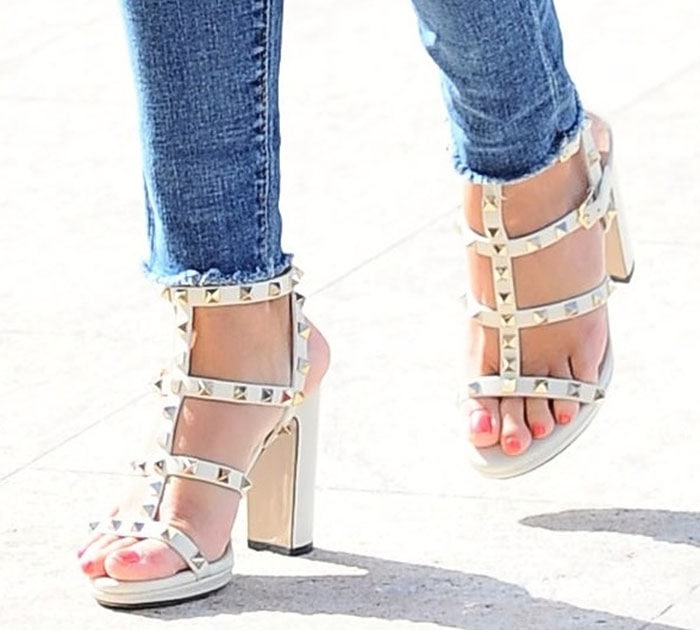 Reese Witherspoon’s white Valentino Rockstud sandals create a leg-lengthening effect with their nude-toned straps, open-toe design, and tall block heels that add height without sacrificing comfort