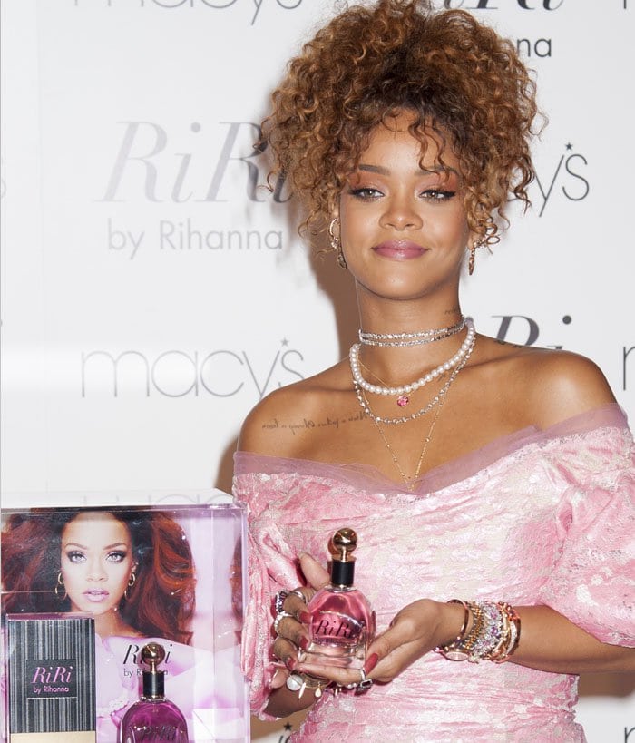 Rihanna smiles and flaunts a bottle of "RiRi by Rihanna" while showing off her stacks of bracelets and layered necklaces