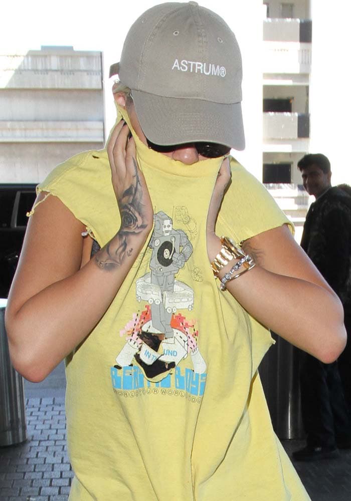Rita covers her hair with a baseball hat and pulls her muscle tee up over her face as she walks through London Heathrow