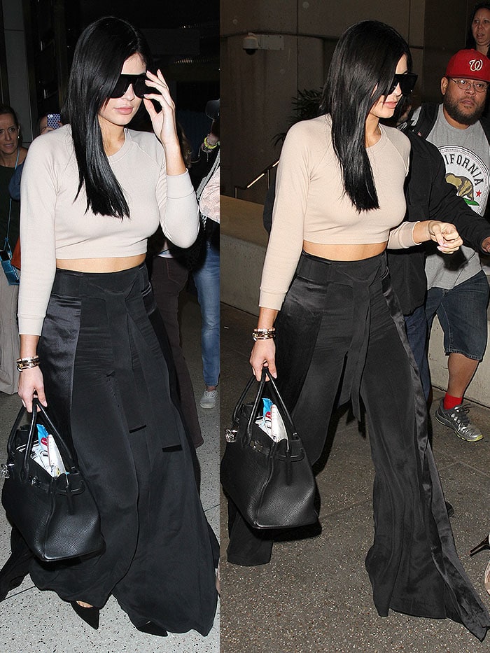 Kylie Jenner wearing a beige crop top and black high-waisted wide-leg pants accessorized with oversized sunglasses, a Hermes Birkin bag, and Gianvito Rossi suede pumps