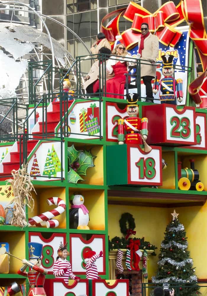 Mariah Carey is seated on an advent calendar-themed float, surrounded by festive decorations and performers, as she leads the Macy's Thanksgiving Day Parade in New York