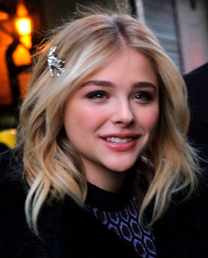 Chloë Grace Moretz looks chic with her blonde hair styled in loose waves, accessorized with a stylish hair clip, while wearing a patterned top and a cozy black coat