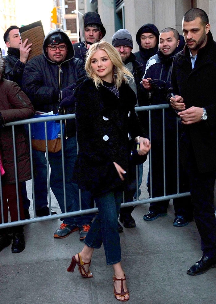Chloë Grace Moretz wears a black coat over a high-neck top and a pair of three-quarter length jeans