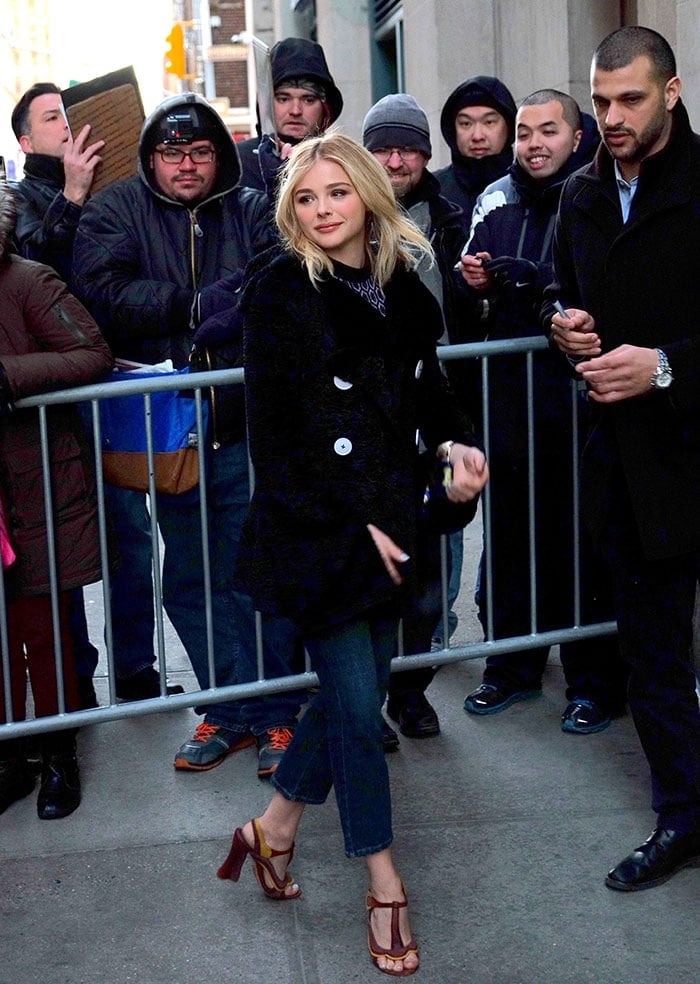 Chloe Grace Moretz wears a black coat over a high-neck top and a pair of three-quarter length jeans