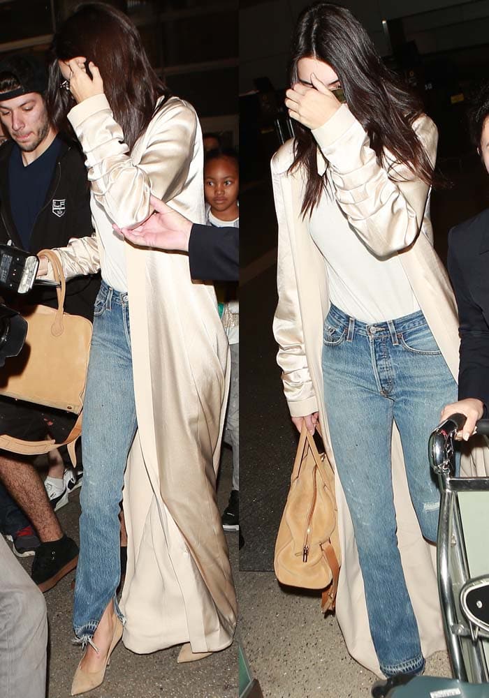 Kendall Jenner steps out in a chic, casual ensemble featuring a silky beige duster, classic blue jeans, a white tee, and nude pointed-toe heels, accessorized with a tan suede handbag