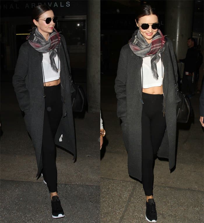 Miranda Kerr in a long dark grey coat paired with a white crop top, black leggings, black sneakers, and a patterned scarf, accessorized with sunglasses and a black handbag