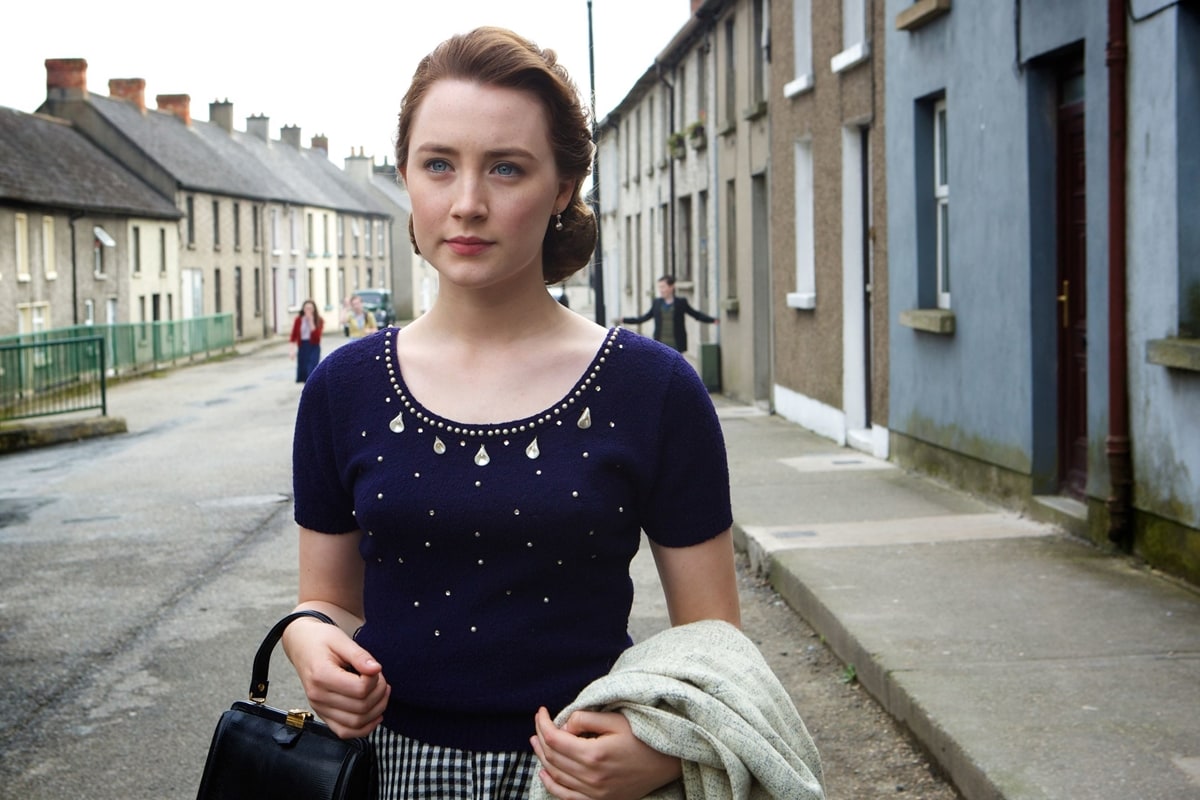 Saoirse Ronan as Eilis Lacey wears a vintage-inspired navy sweater with jeweled detailing and a gingham skirt, capturing 1950s Irish fashion in Brooklyn