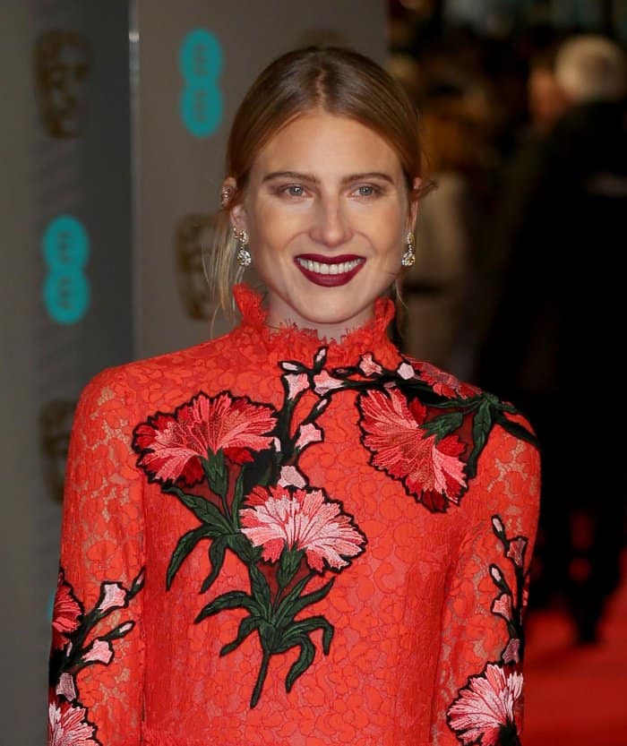 Dree Hemingway brightens the red carpet in a bold high-neck lace gown embroidered with vibrant floral appliqués, pairing the statement look with deep berry lipstick and sparkling earrings