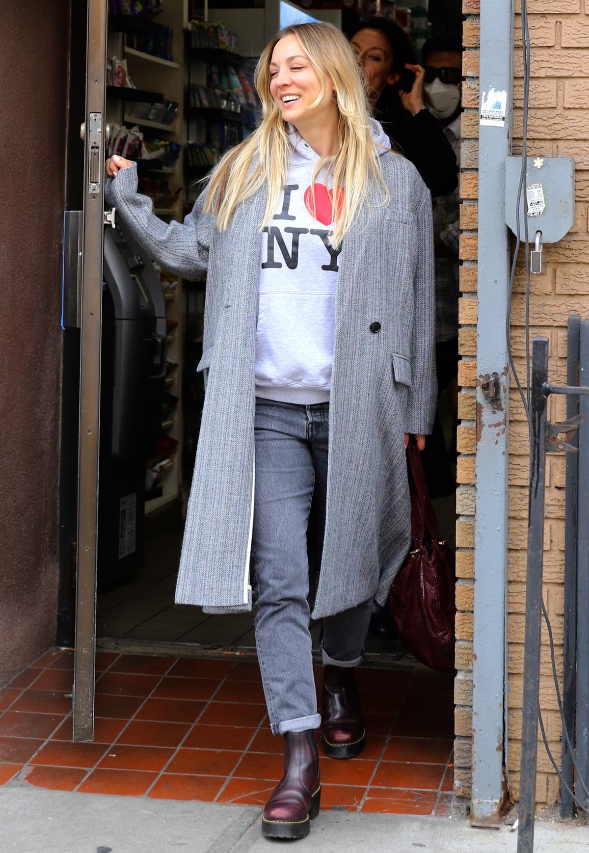 Kaley Cuoco wears an 'I Love New York' hoodie with a grey overcoat, cuffed faded denim jeans, and boots with the famous yellow Dr. Martens stitch while filming The Flight Attendant