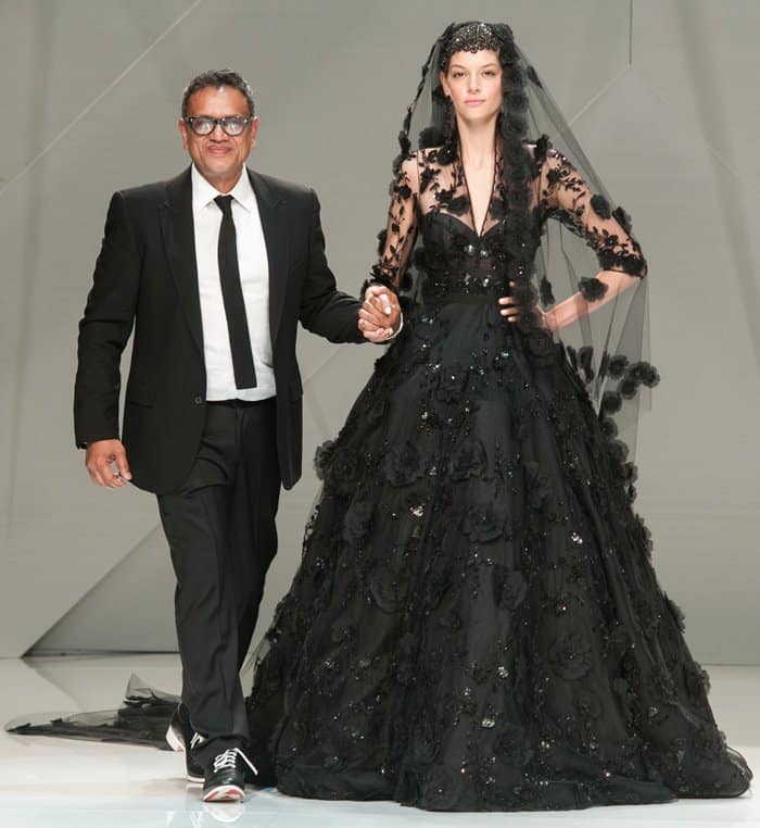 Naeem Khan takes a bow with a model showcasing a dramatic black bridal gown adorned with intricate floral details at Pasarela Costura España