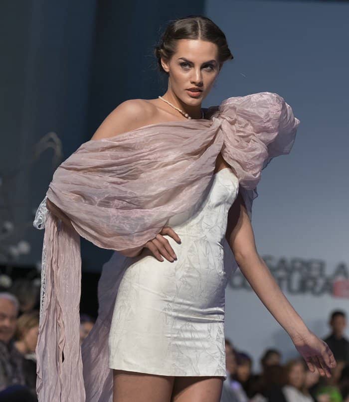 A model showcases an asymmetrical white mini dress with a sheer shawl at Pasarela Costura España