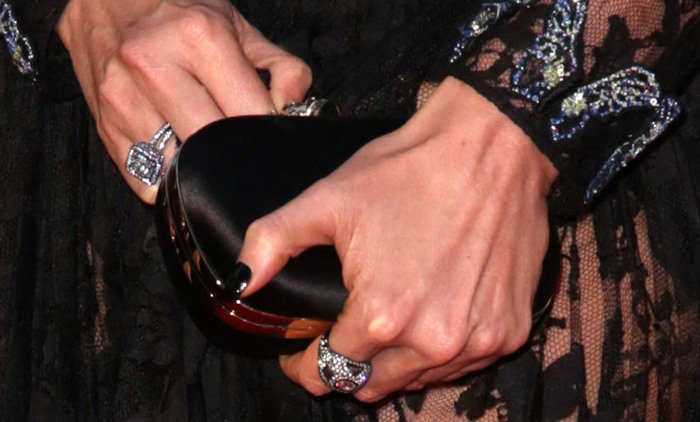 Charlize Theron accessorizes her stunning look with a sleek black Alexander McQueen clutch, complemented by striking statement rings that add a touch of opulence to her ensemble