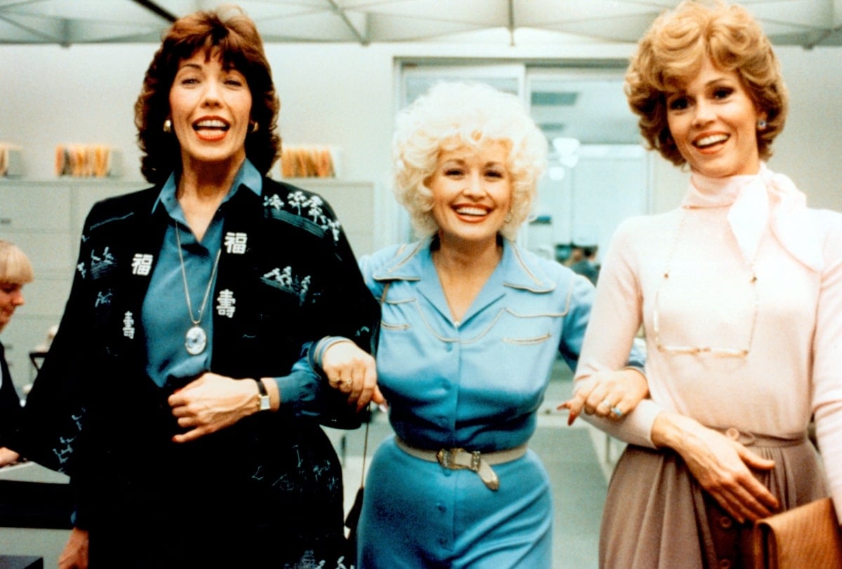 Lily Tomlin, Dolly Parton, and Jane Fonda exude retro office-chic in 9 to 5 (1980), with Tomlin in a bold embroidered jacket, Parton in a Western-inspired belted blue dress, and Fonda in a soft pink blouse with a high-waisted skirt, capturing the film’s empowering style