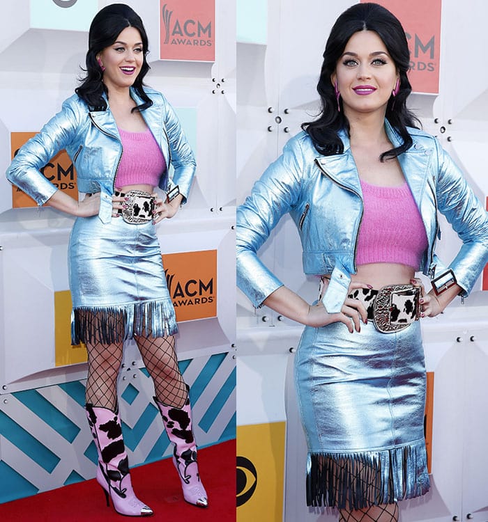 Katy Perry wears a metallic blue jacket and fringed skirt with a pink crop top and pink cow-patterned cowboy boots