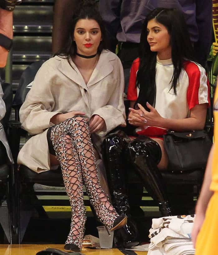 Kendall Jenner turns heads at a Lakers game in eye-catching lace-up boots paired with an oversized coat, while sister Kylie opts for a sleek patent leather look