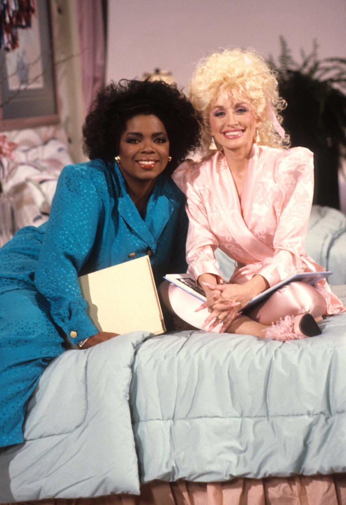 Oprah Winfrey and Dolly Parton share a lighthearted moment on the set of Dolly in 1987, with Oprah looking elegant in a teal double-breasted suit, while Dolly stuns in a silky pink wrap ensemble with matching satin pants and playful feather-trimmed heels
