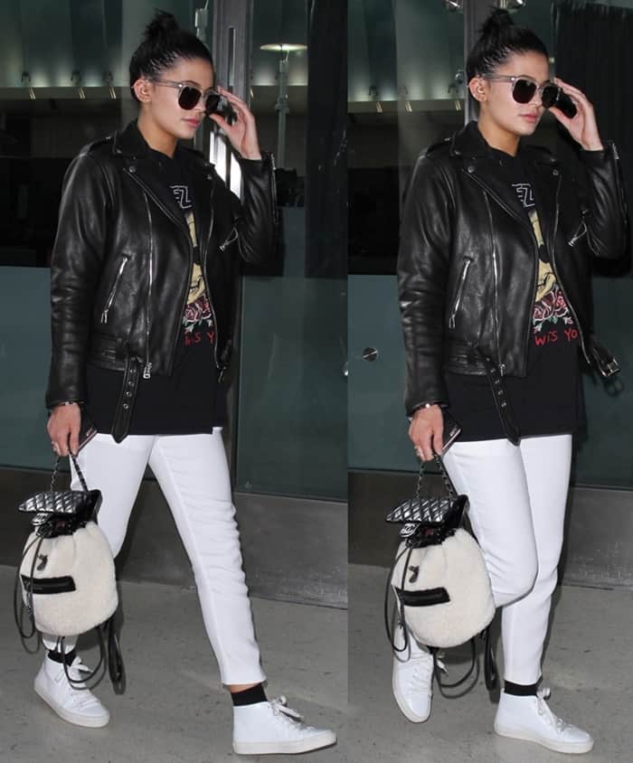 Kylie Jenner keeps it effortlessly edgy at LAX, pairing a black leather biker jacket with a Yeezus tour tee, white pants, classic white sneakers, and a cozy Chanel shearling backpack