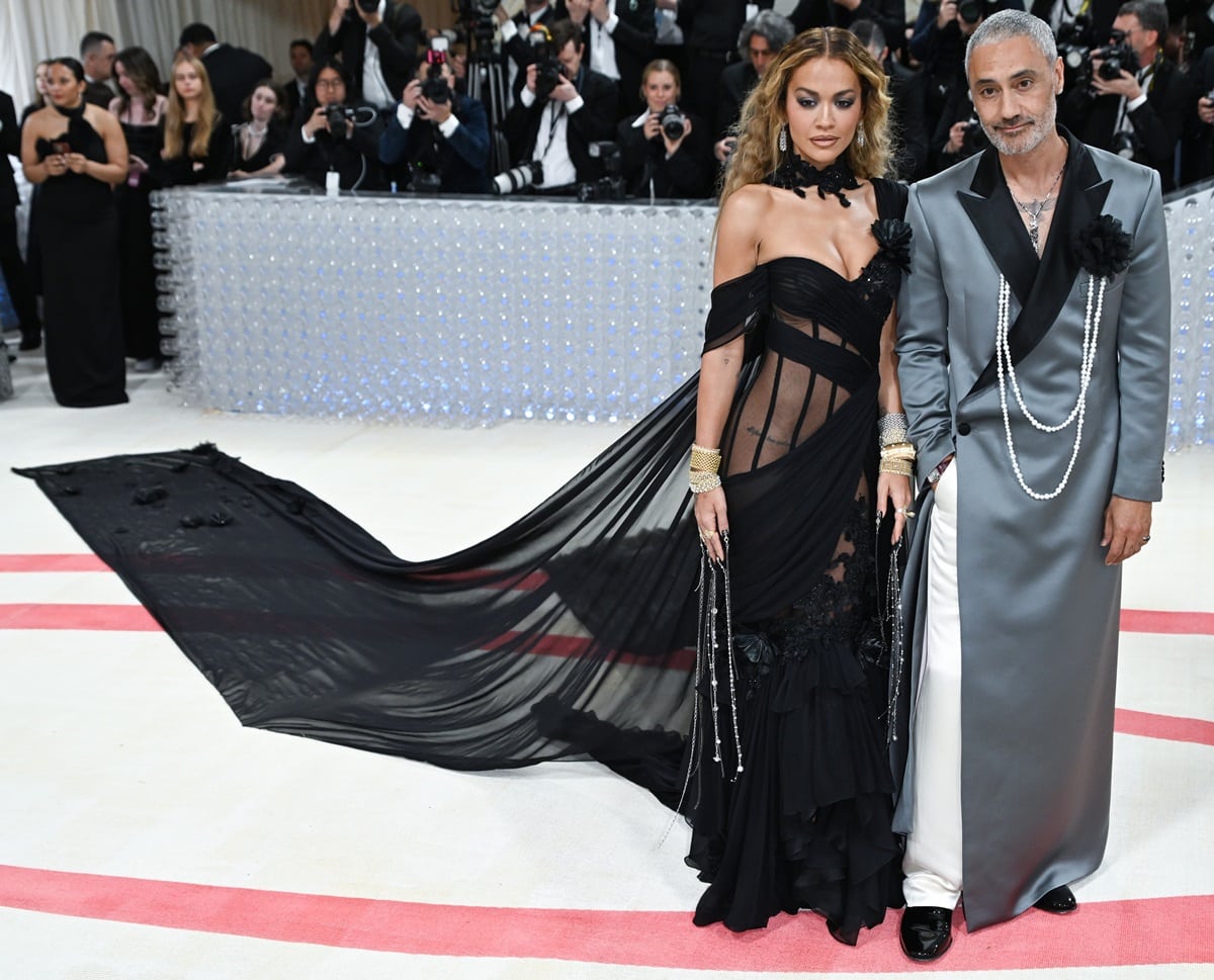 Rita Ora arrived at the 2023 Met Gala in a sheer black gown with a dramatic train and a unique manicure, while her husband, Taika Waititi, coordinated with her look by wearing extra-long strands of pearls