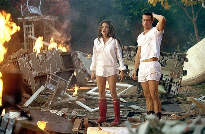 Angelina Jolie and Brad Pitt capture a pivotal moment in their 2005 film, "Mr. and Mrs. Smith," where Angie, clad in a white men's shirt and striking red Hunter rain boots, stands beside Brad in an intense scene amidst the destruction of their on-screen home