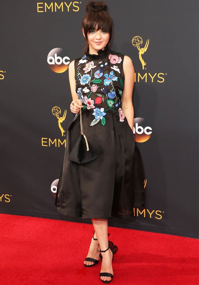 Maisie Williams stuns in a floral-embroidered black midi dress paired with strappy black heels, a sleek updo, and bold green eye makeup, creating a chic yet edgy red-carpet look at the Emmys