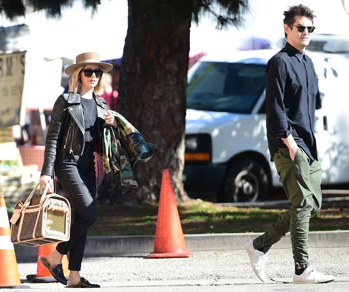 Ashley Tisdale pairs a black leather jacket with pinstripe pants and sleek black loafers, adding a beige fedora and pet carrier for a chic yet casual look, while Christopher French keeps it relaxed in a black button-up, olive-green joggers, and white sneakers at the Los Angeles Farmers Market on January 27, 2019