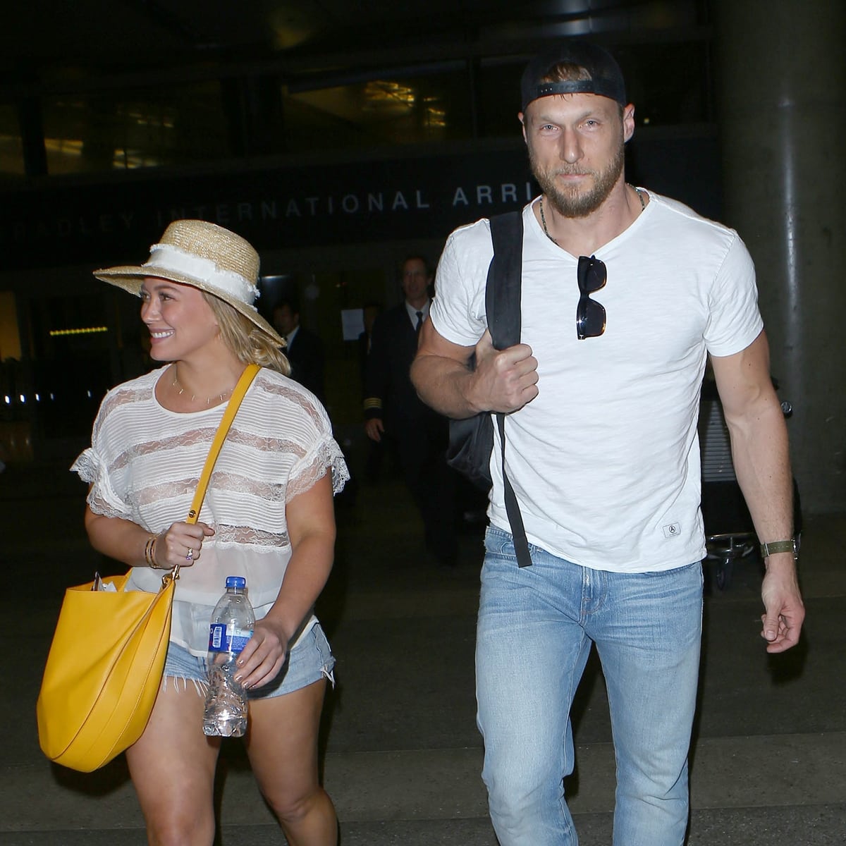 Hilary Duff keeps it effortlessly chic in a sheer lace top, frayed denim shorts, and a straw hat, while Jason Walsh keeps it casual in a white tee and jeans at LAX
