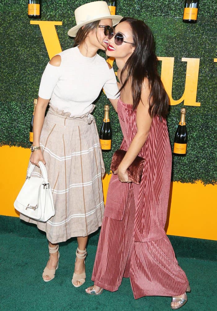 Jamie Chung looks chic in a cold-shoulder top and pleated midi skirt, while Cara Santana stuns in a pink pleated jumpsuit as the two share a playful moment at the Veuve Clicquot Polo Classic