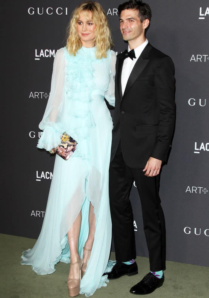Brie Larson stuns in a flowing mint Gucci gown paired with nude platform heels and a colorful clutch, while Alex Greenwald complements her look in a classic black tuxedo with a pop of color from his vibrant socks