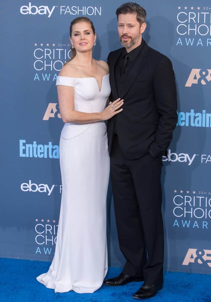 Amy Adams stuns in a white off-the-shoulder Atelier Versace gown, paired with Jimmy Choo shoes and sparkling jewelry, as she poses alongside her husband, Darren Le Gallo