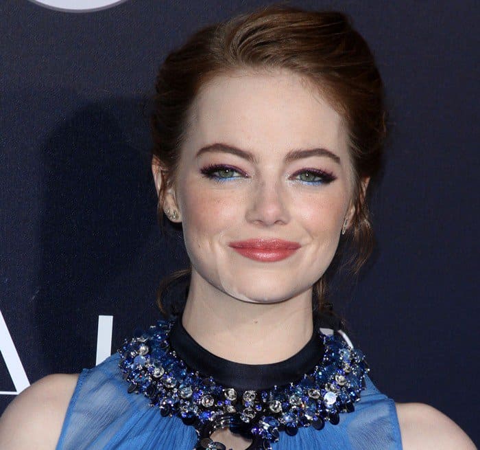 Emma Stone stuns with a sophisticated updo, bold winged eyeliner, shimmering blue eyeshadow, and a soft pink lip, perfectly complementing her embellished neckline