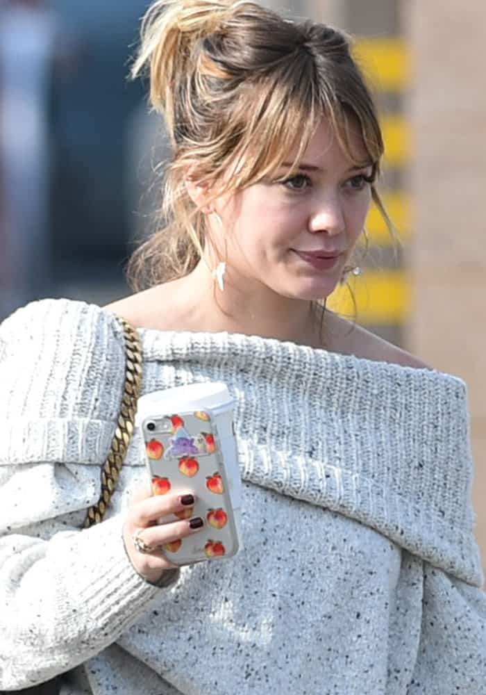Hilary Duff steps out in a cozy off-shoulder speckled sweater while carrying a phone with a playful peach-themed case and a to-go coffee cup