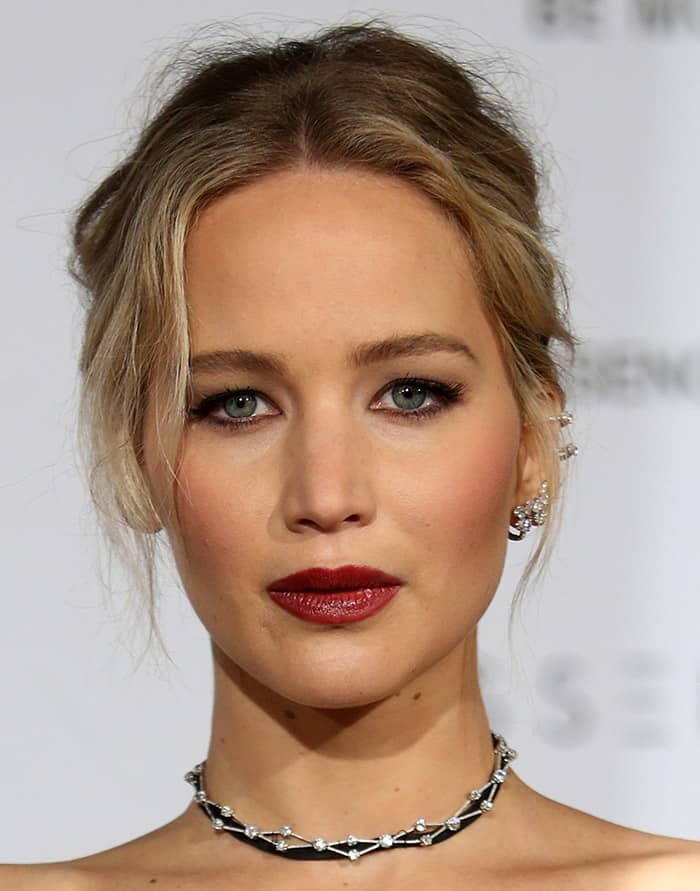 Jennifer Lawrence enhances her look with striking dark red lips, bold black eyeliner, and subtle eyeshadow that complements her blue-gray eyes