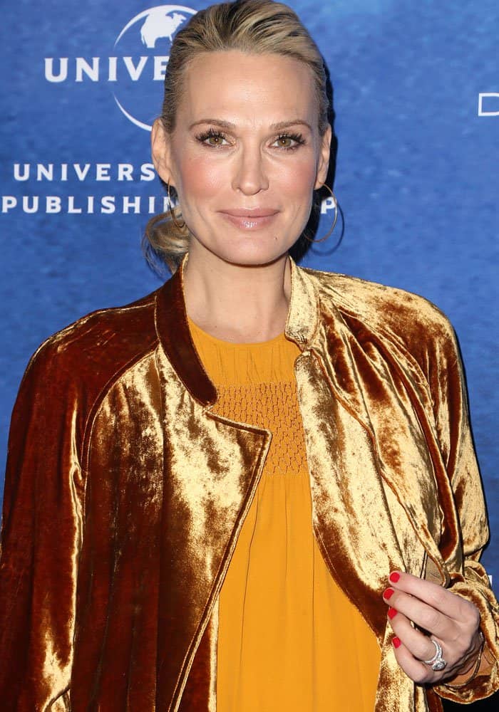 Molly Sims exudes elegance with a sleek low bun, natural glowing makeup, and delicate hoop earrings, perfectly complementing her golden velvet coat